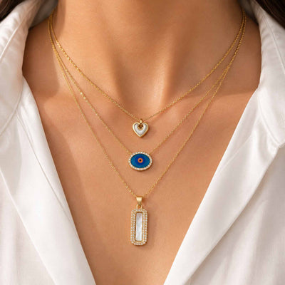 Gold necklace stack featuring a mother-of-pearl heart pendant, blue evil eye pendant, and rectangular mother-of-pearl pendant styled on a model.