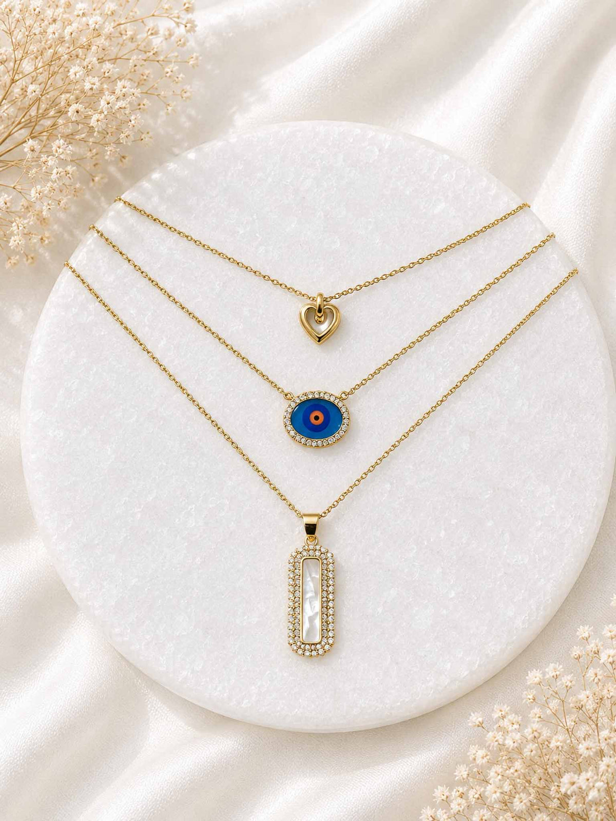 Three-layer gold necklace stack with heart, blue evil eye, and rectangular mother-of-pearl pendants displayed on a white stone surface.