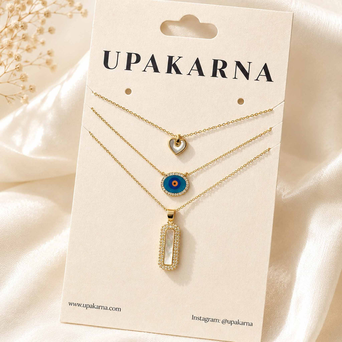 UPAKARNA gold necklace stack featuring a mother-of-pearl heart, blue evil eye pendant, and rectangular pearl pendant displayed on a branded jewellery card.