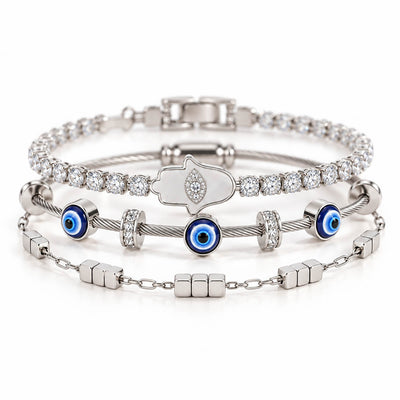 A product photo of a stacked set of silver-tone bracelets featuring a crystal tennis chain with a mother-of-pearl Hamsa hand pendant, a twisted cable wire band with blue evil eye beads, and a link chain with small cube charms.