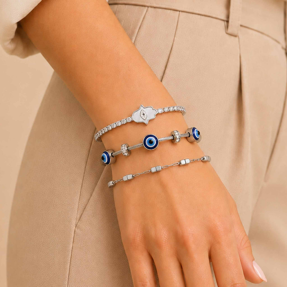 Close-up of a wrist wearing a silver bracelet stack featuring a sparkling Hamsa tennis bracelet, a blue Evil Eye bangle with silver accents, and a minimalist geometric chain bracelet, styled with a neutral beige outfit.