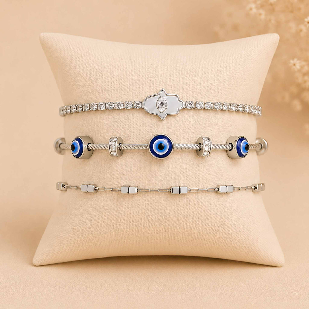 Silver bracelet stack displayed on a cream cushion, featuring a sparkling Hamsa bracelet, a blue Evil Eye bracelet with silver accents, and a minimalist silver chain bracelet.