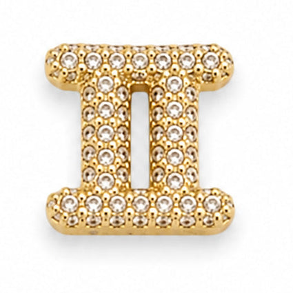 Gold-tone Gemini zodiac charm featuring the distinctive Roman numeral-style twin symbol, with two vertical columns connected by horizontal bars. The charm is densely embellished with numerous small white rhinestones, creating a sparkling textured surface. It has polished gold-tone edges, raised detailing, and a dimensional finish.