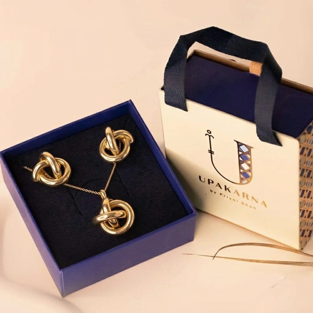 A premium gold-tone jewelry set displayed in an open navy blue box, featuring a necklace and matching stud earrings with interlinked circular knot designs. The set is paired with an elegant branded Upakarna gift bag in cream and navy, placed on a neutral beige surface, highlighting a refined and luxurious presentation.