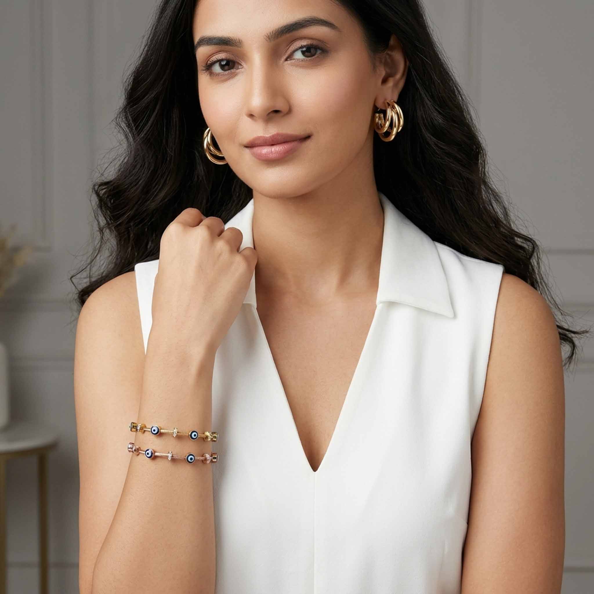 A woman wearing a sleeveless white top models a pair of gold triple-hoop earrings and two evil eye charm bracelets in gold and rose gold. The bracelets feature blue evil eye beads and crystal accents, creating a modern, elegant look against a neutral indoor background.