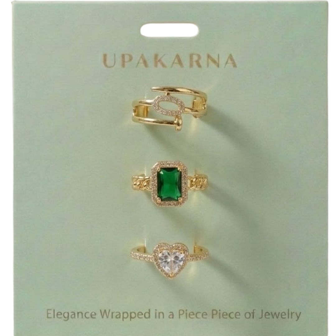 A stack of three adjustable gold rings featuring a heart-shaped crystal, an emerald-green stone, and a sparkling oval nail-head band.