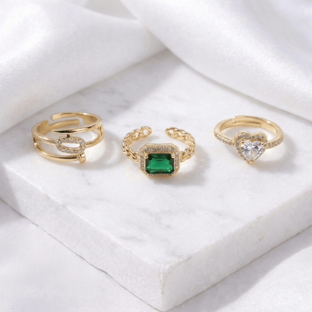 A stack of three adjustable gold rings featuring a heart-shaped crystal, an emerald-green stone, and a sparkling oval nail-head band.