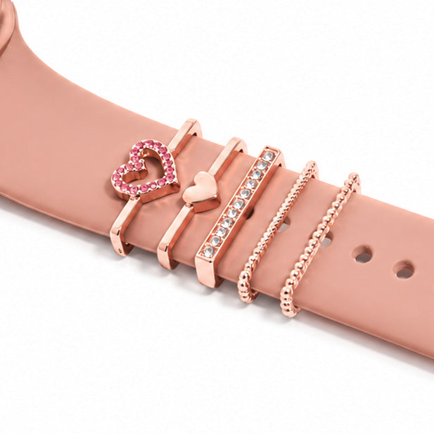 Rose gold smartwatch strap close-up decorated with a pink crystal heart charm, heart charm, pavé crystal bar, and delicate chain-style bands on a white background.
