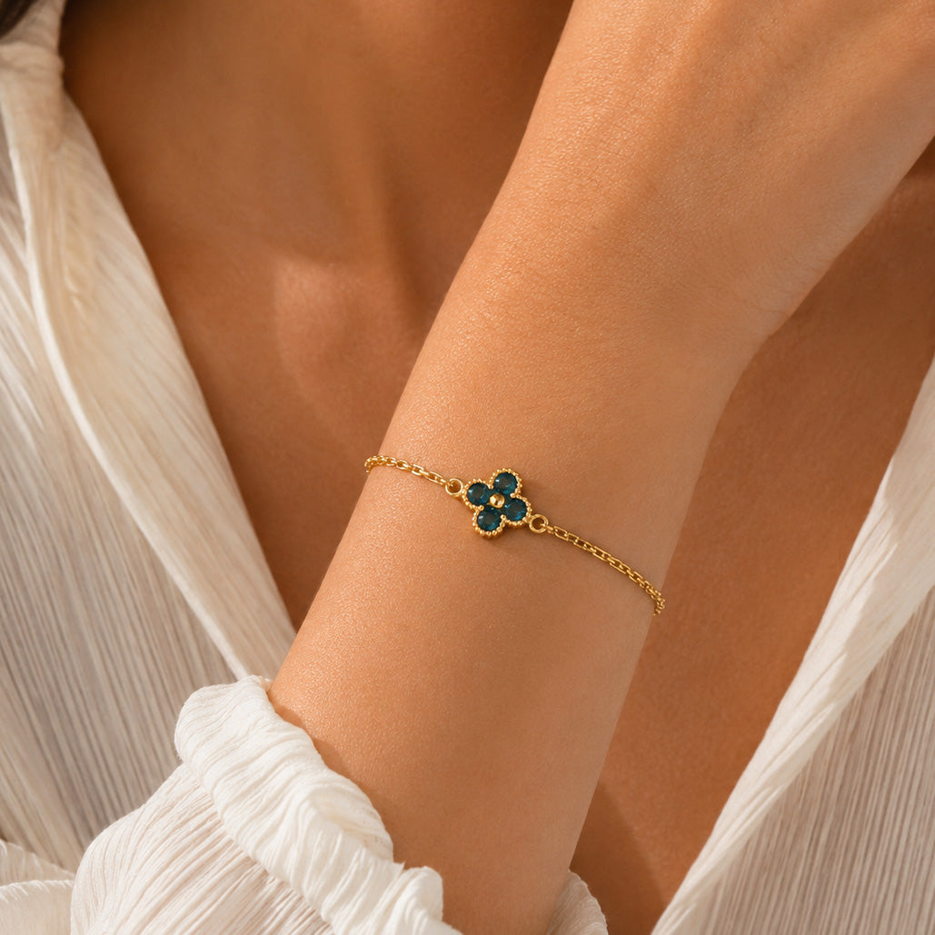 Gold-plated bracelet featuring a delicate four-leaf clover charm with deep teal-blue stones, worn on the wrist with a white outfit in warm natural sunlight.