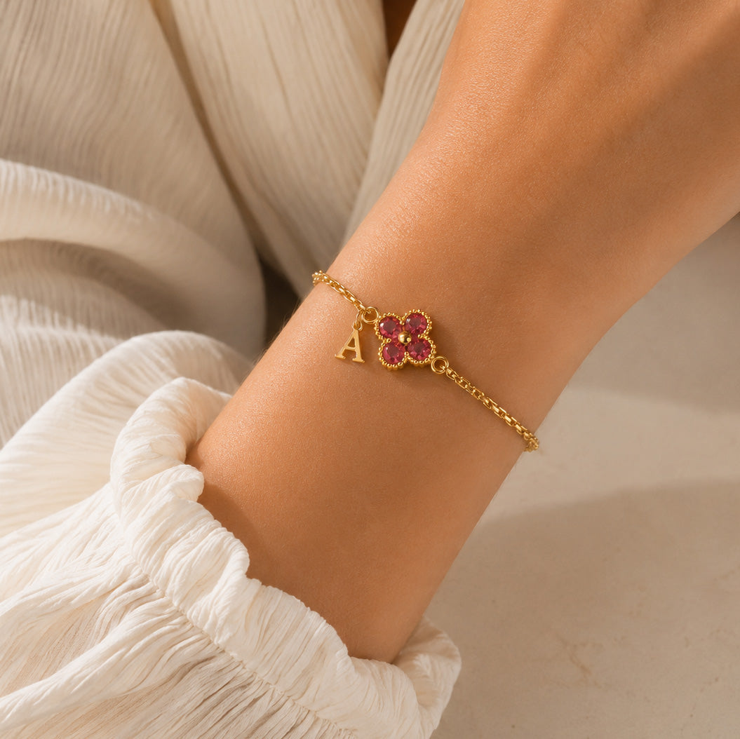 Gold-plated personalized bracelet featuring a delicate pink four-leaf clover charm with an “A” initial, worn on the wrist with a white outfit in warm natural sunlight.