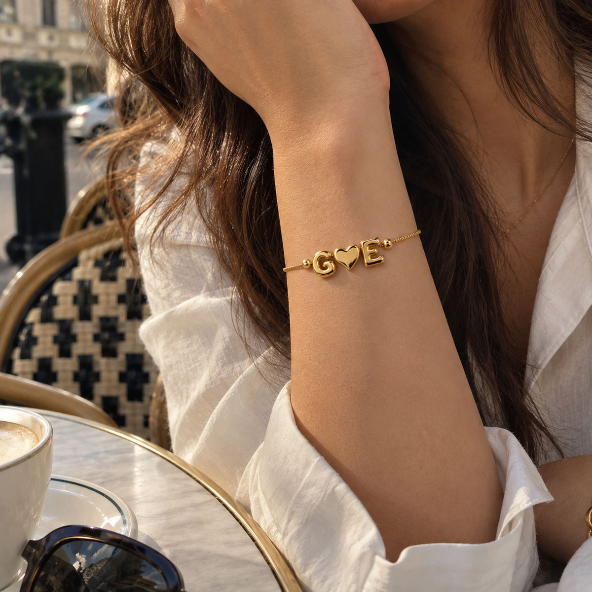 Lifestyle close-up of a woman wearing a delicate gold-tone personalized bracelet with G♥E charms, styled with a white outfit at an outdoor café.