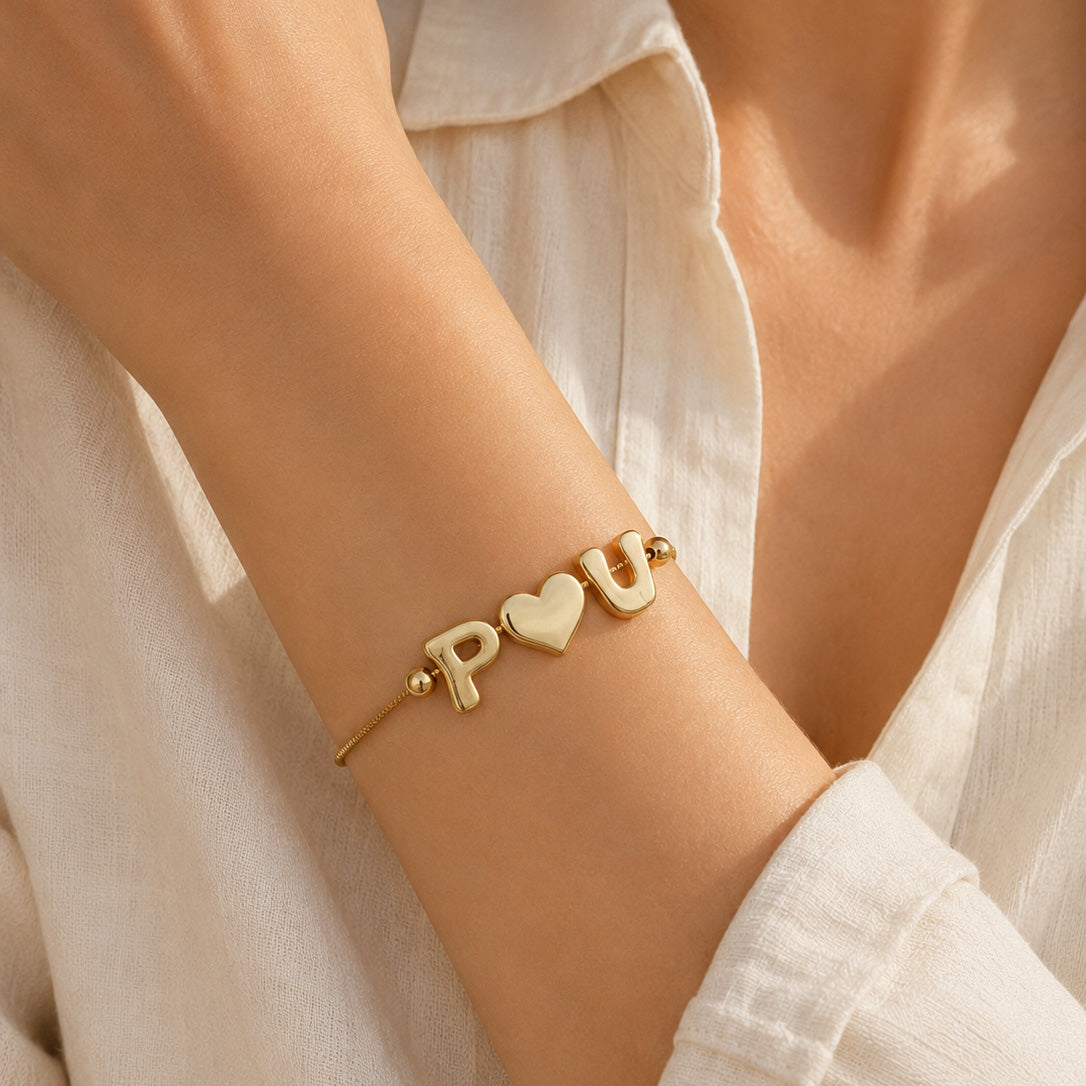Gold-plated delicate bracelet worn on a model, featuring P♥U initial-and-heart charms, styled with a white outfit in warm natural sunlight.