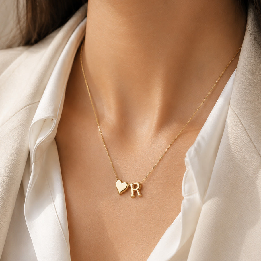 Gold-plated delicate chain necklace worn on a model, featuring a polished ♥R heart-and-initial pendant, styled with a white outfit in warm natural sunlight.
