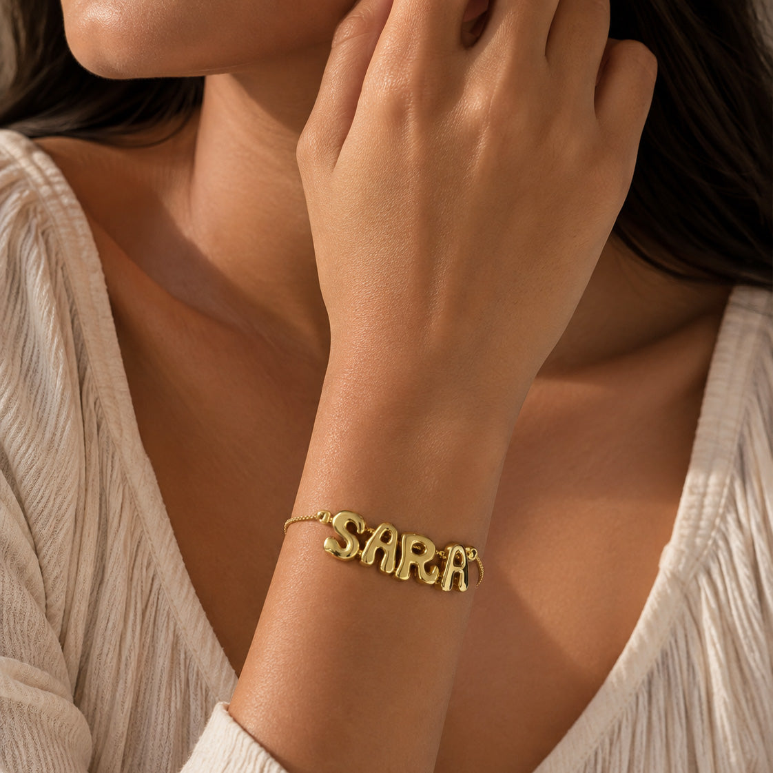 Gold-plated personalized bracelet featuring the name “SARA” on a delicate chain, worn on the wrist in a warm, natural-light close-up with a cream textured outfit.