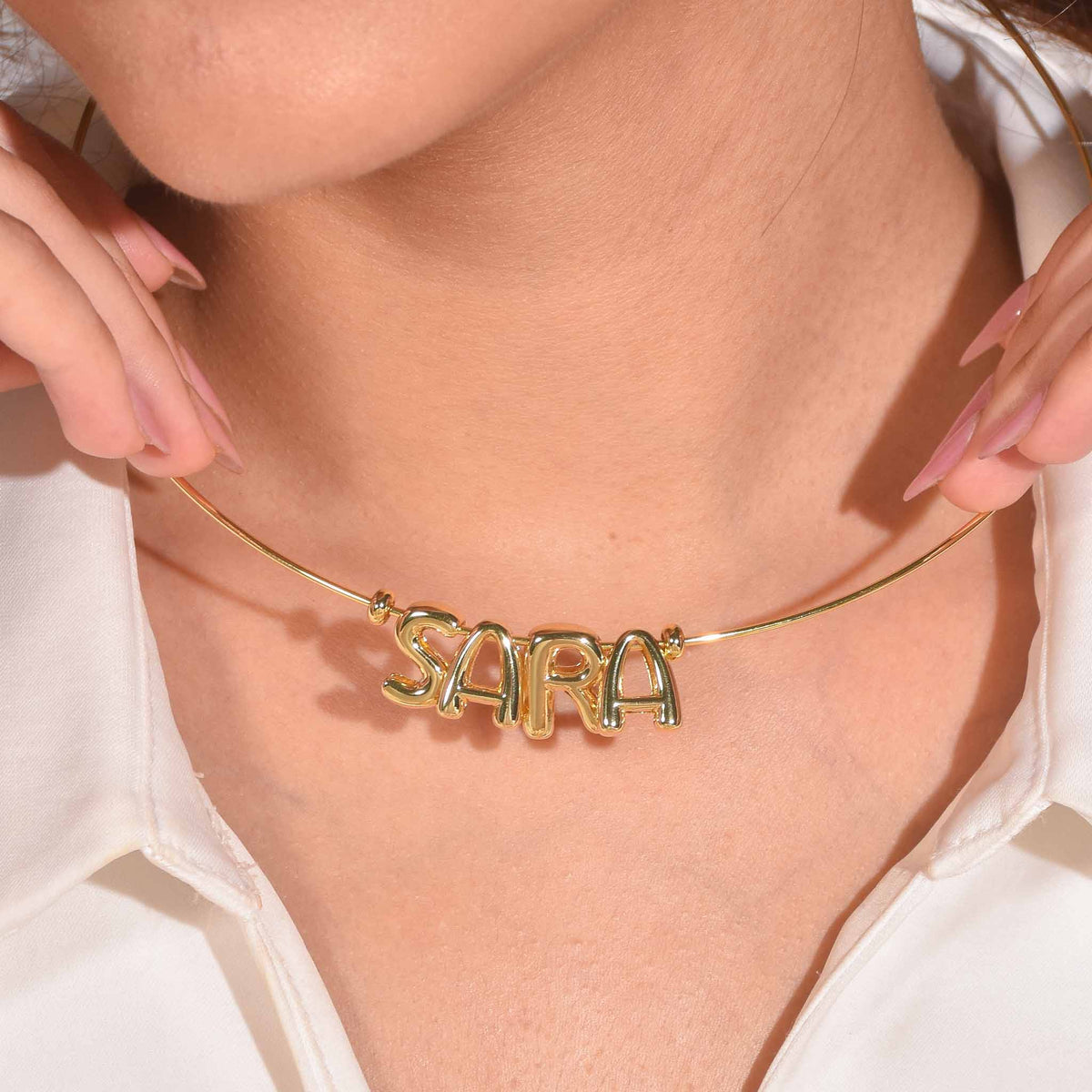 Gold tone name necklace worn around the neck, featuring the name “SARA” on a delicate chain, styled with a white outfit.