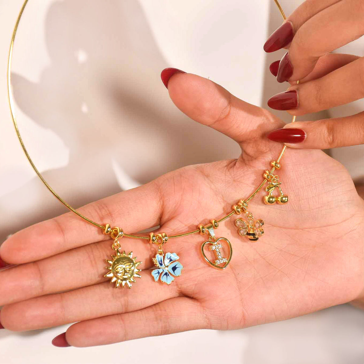 Gold-tone charm necklace displayed across an open hand, featuring sun, blue flower, heart, and other dangling charms on a delicate chain.