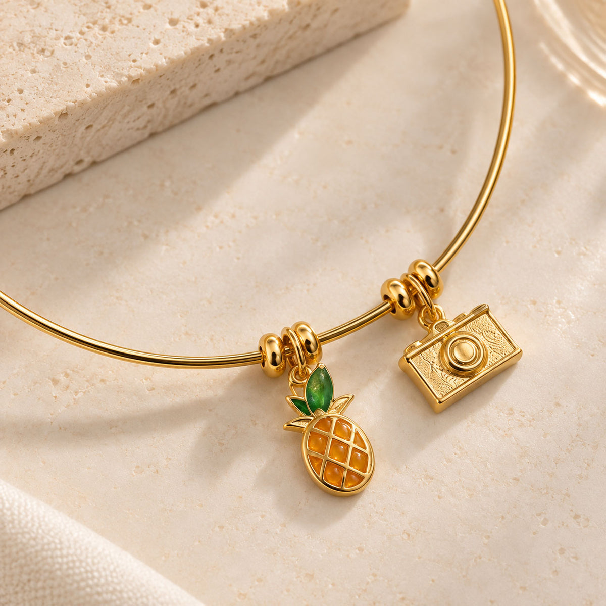 Gold-tone charm necklace close-up on a beige stone surface, featuring pineapple and camera shaped charms with a delicate gold chain.