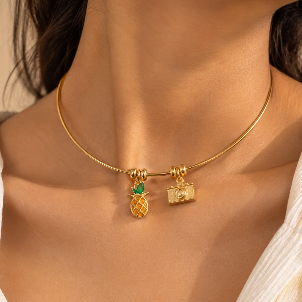 Gold-tone charm necklace worn around the neck, featuring pineapple and camera charms on a delicate chain, styled with a white outfit.