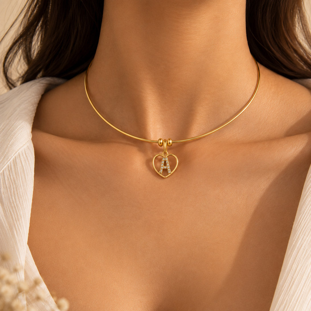 Gold-tone necklace worn around the neck, featuring a heart-shaped pendant with crystal detailing on a delicate chain, styled with a white outfit.