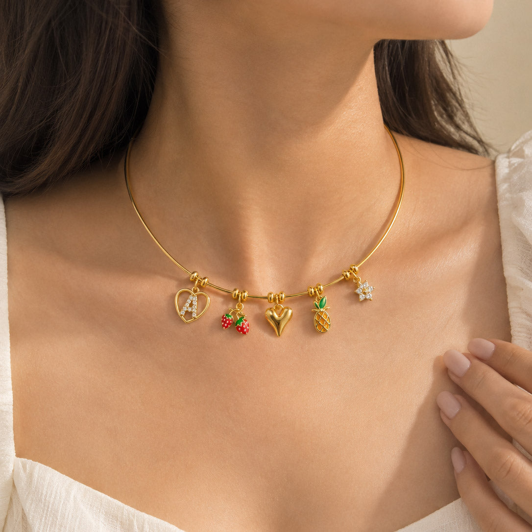 Gold-tone charm necklace worn with a white outfit, featuring multiple decorative pendants including a heart-shaped initial, red fruit charms, a heart charm, a pineapple charm, and crystal detailing.