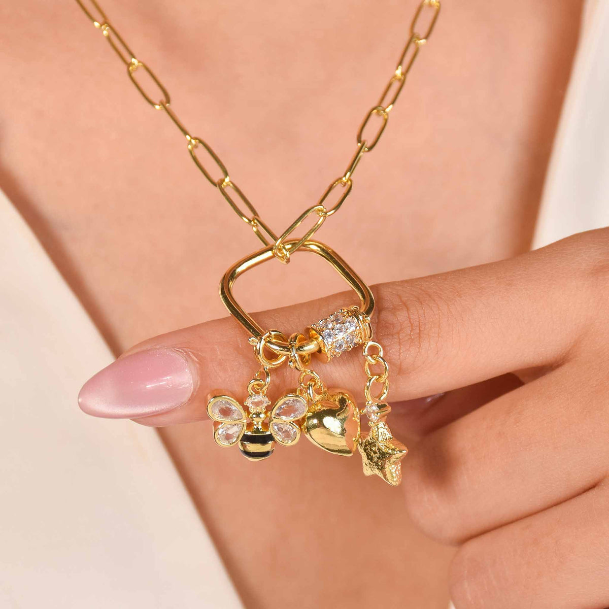 Gold-tone charm necklace with a square charm holder featuring bee, heart, and star charms, worn against a neutral outfit.