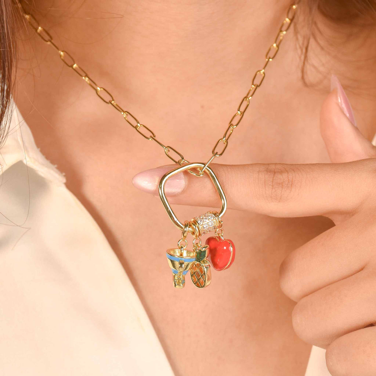 Gold tone charm necklace featuring a rectangular charm holder with a crystal-studded clasp and colorful dangling charms, including a blue cup, pineapple, and red heart, worn on a model.