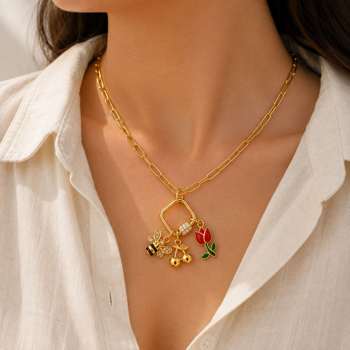 Gold-tone charm necklace featuring a rectangular charm holder with a crystal-studded clasp, decorated with a bee, red tulip, and dangling gold bead charms, worn with a white outfit.
