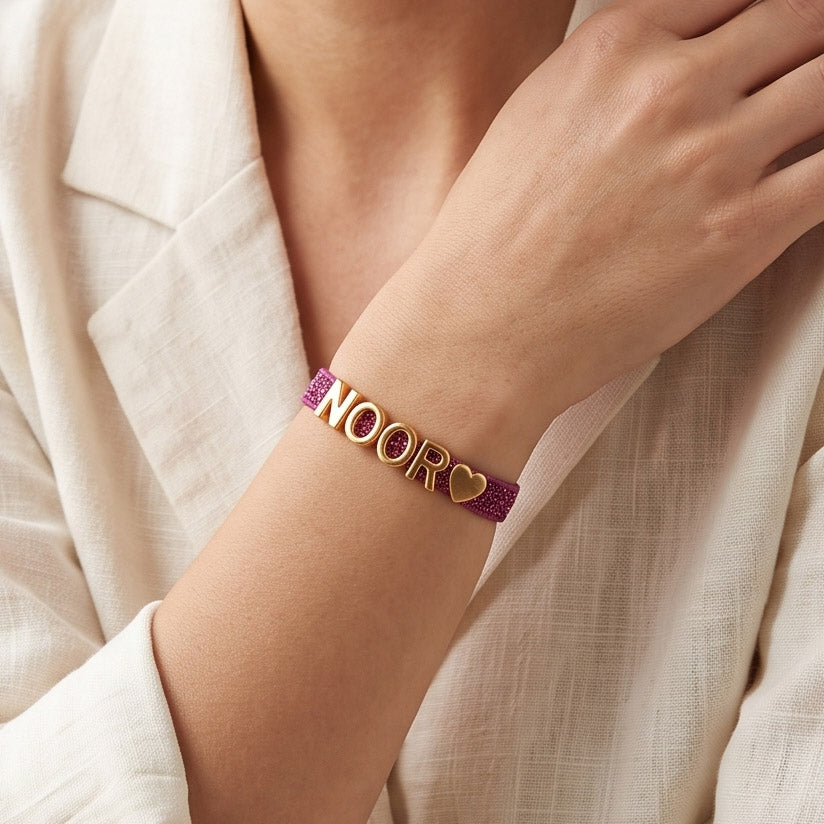 Lifestyle close-up of a hand wearing a pink glittery personalized bracelet with gold-tone NOOR letter charms and a heart charm, styled with a white linen outfit.
