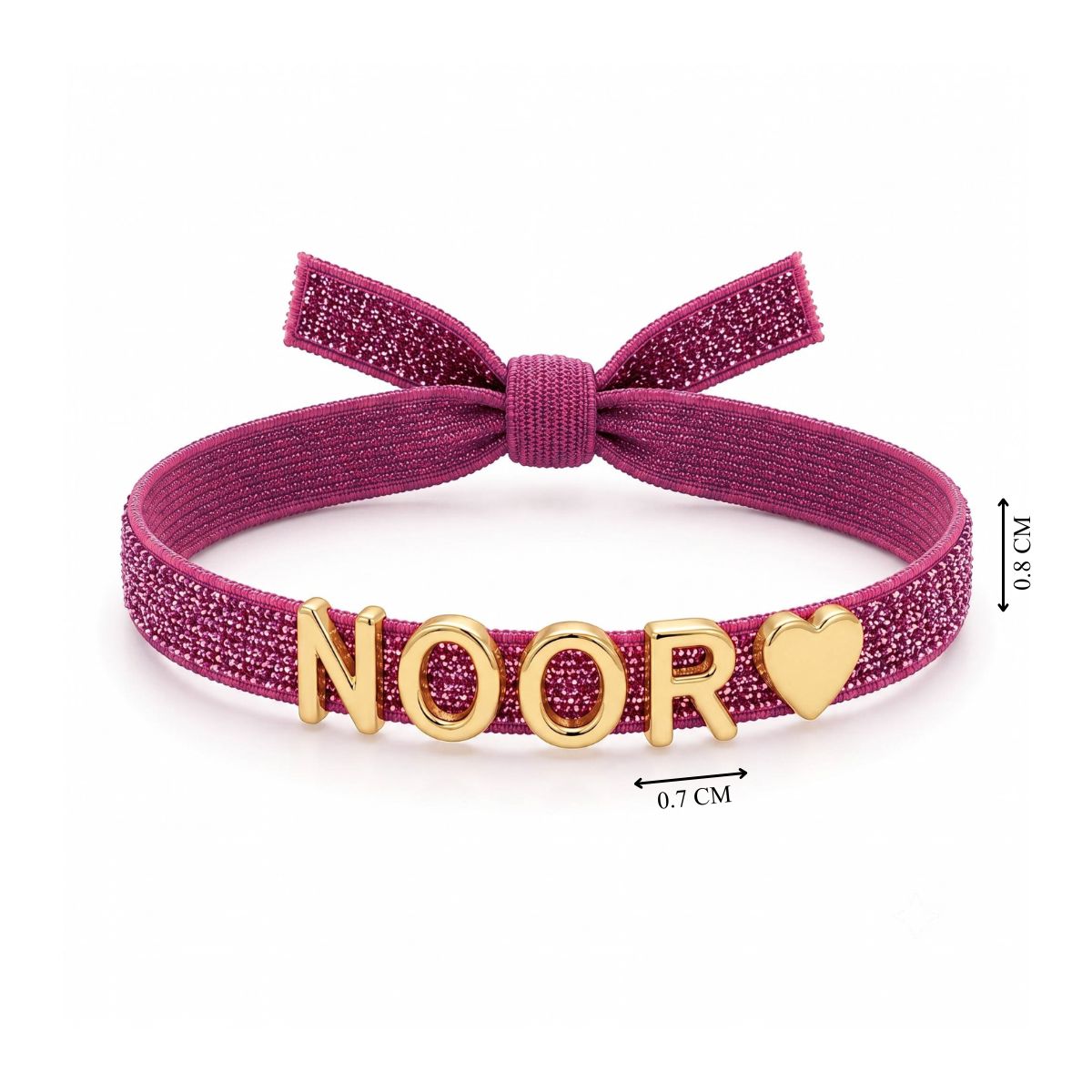 Pink glittery adjustable bracelet featuring a bow at the top and gold-tone NOOR letter charms with a heart charm at the end. The bracelet band is approximately 0.8 cm wide.