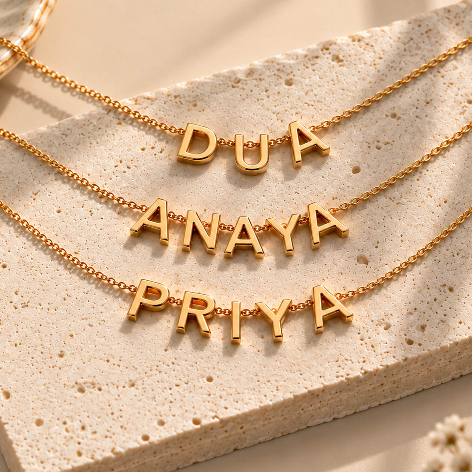 A flat-lay product photo of three dainty gold name necklaces arranged neatly on a textured, light beige stone slab, displaying customized letters that spell "DUA", "ANAYA", and "PRIYA".