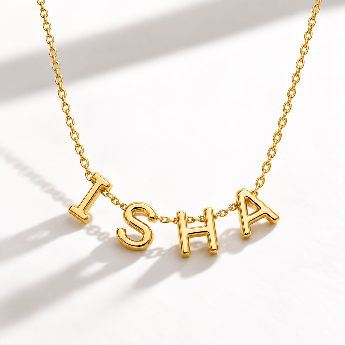 A studio flat-lay photo of a thin, dainty gold link necklace centered with polished gold letter pendants that spell the name "ISHA" against a white background with soft shadows. 