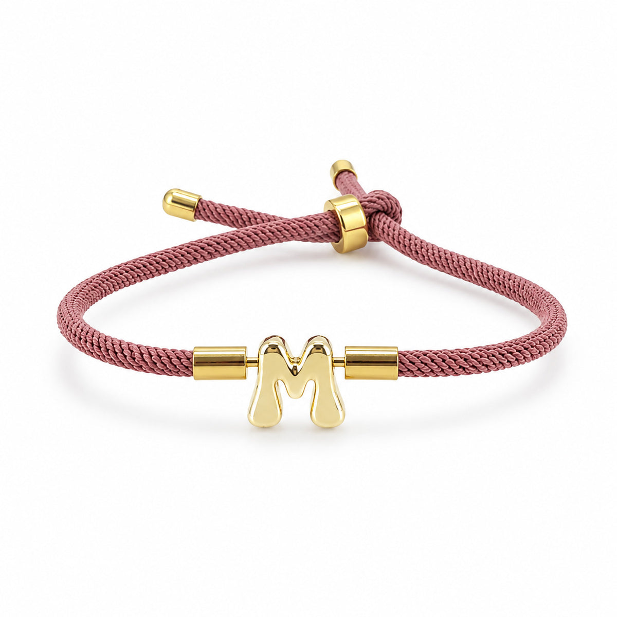 Gold-tone initial “M” charm bracelet with a dusty rose braided cord and adjustable sliding closure, displayed on a clean white background.