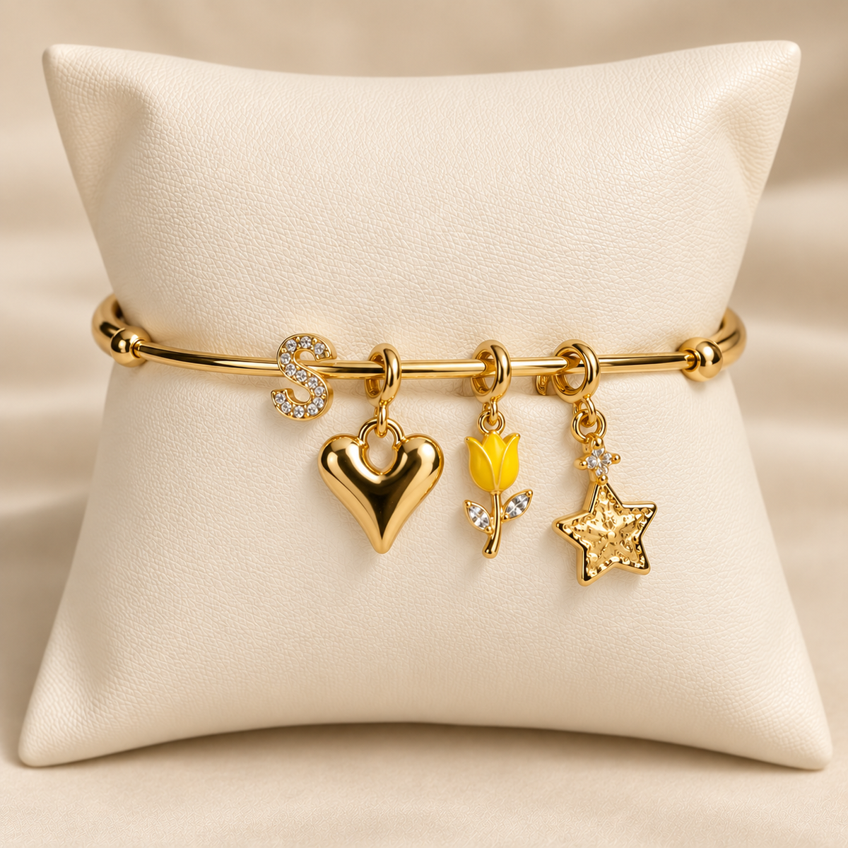 Gold-tone charm bangle displayed on a white cushion, featuring a rhinestone initial “S” charm, a polished gold heart charm, a yellow tulip charm, and a gold star charm.