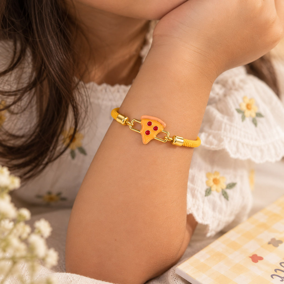 Close-up of a child wearing a yellow braided cord bracelet with a gold-tone pizza-slice charm, styled with a white floral outfit.