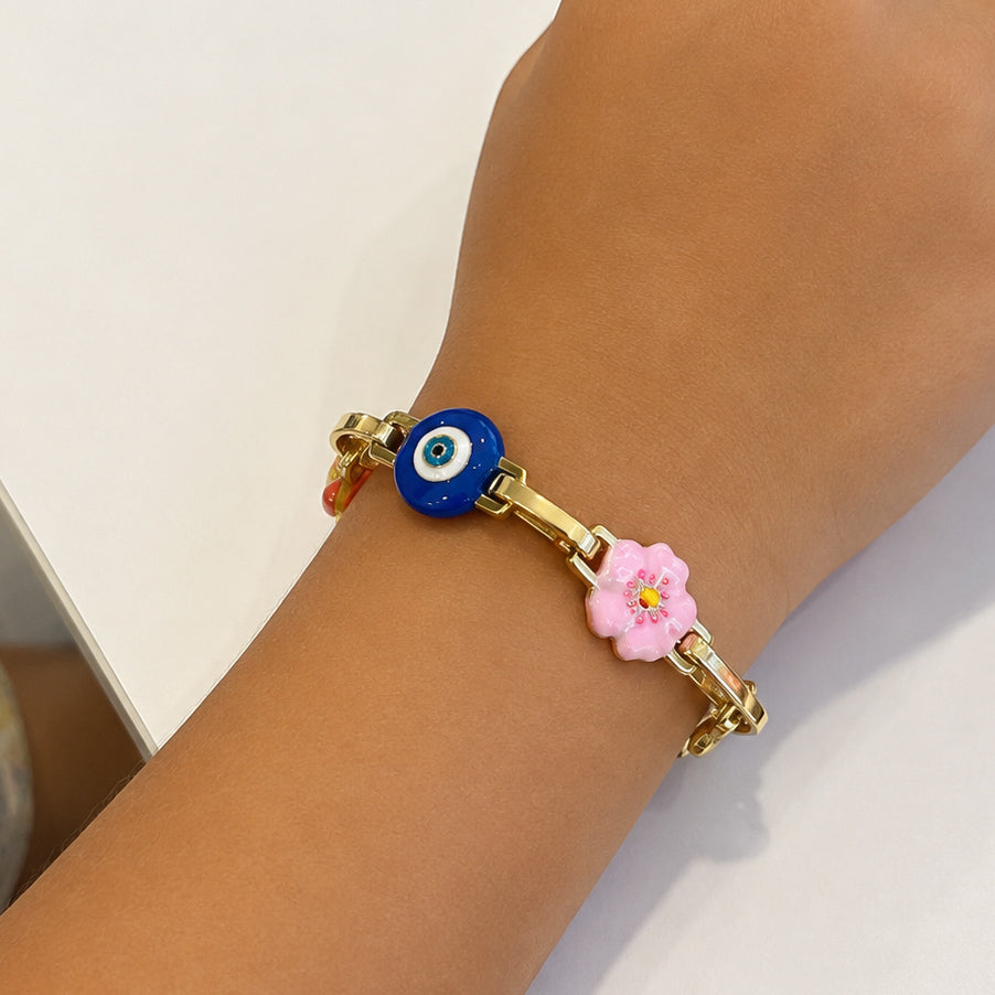 Gold-tone charm bracelet worn on a wrist, featuring a blue evil-eye charm and a pink flower charm with colorful accents.