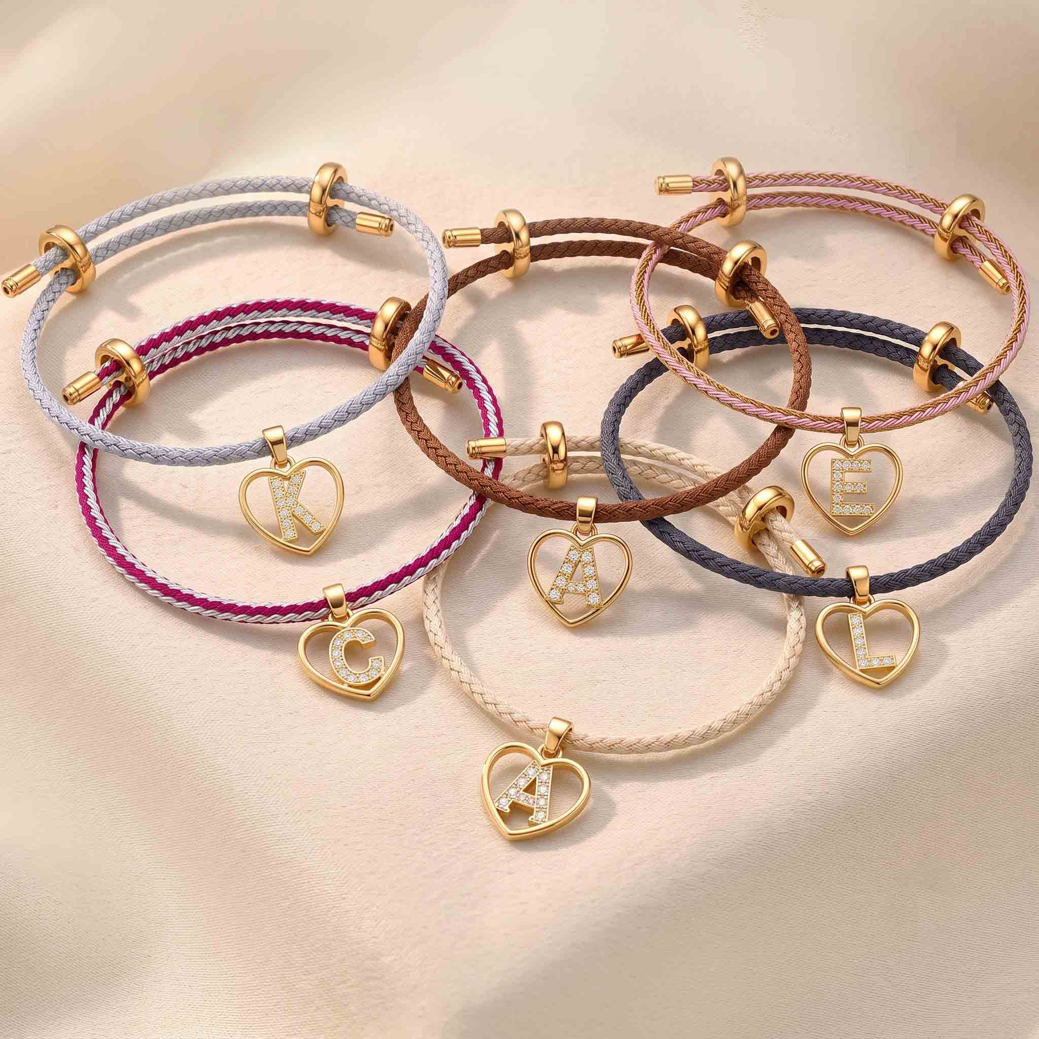 Collection of colourful braided adjustable bracelets featuring gold heart-shaped initial pendants with sparkling letters, displayed together on a soft neutral background.