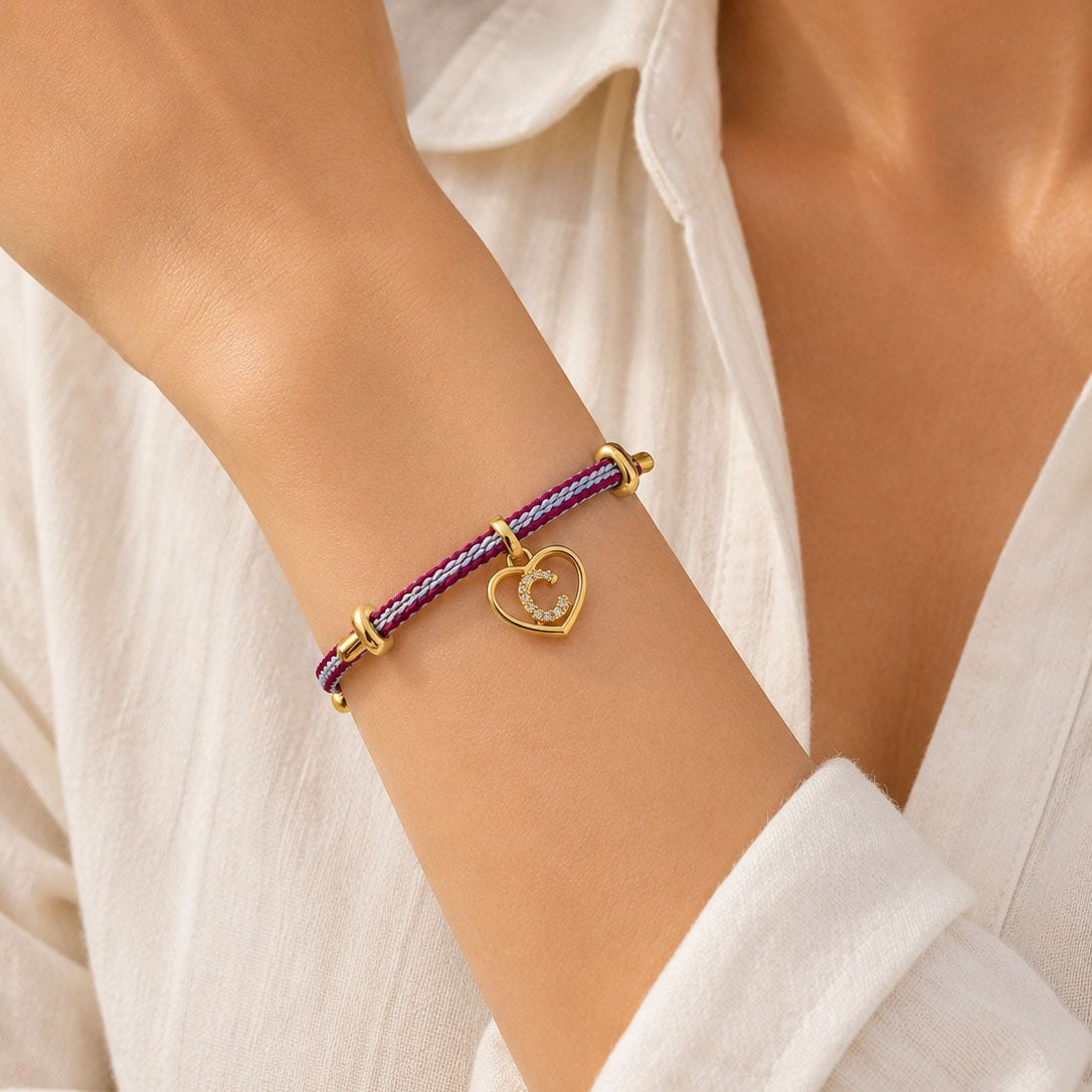 Close-up of a colourful braided bracelet worn on the wrist, featuring a gold heart-shaped pendant with a sparkling letter C initial.
