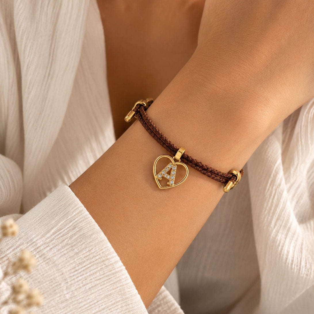 Close-up of a model wearing a brown braided cord bracelet with a gold-tone heart-shaped initial “A” charm embellished with clear crystals, styled with a white textured outfit.