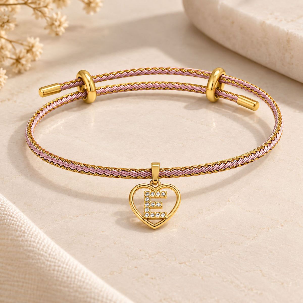 Pink and gold braided cord bracelet with a heart-shaped initial “E” charm embellished with clear crystals, styled on a beige marble surface with soft floral accents.