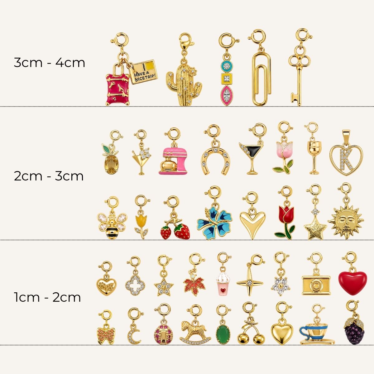 A collection of gold-toned charms with spring-ring clasps organized by size ranges from 1 cm to 4 cm. The assortment features various designs including travel accessories, food and drinks, animals, flowers, celestial symbols, everyday objects, and an uppercase letter "K" charm inside a heart frame, all embellished with colorful enamel and crystal details.   