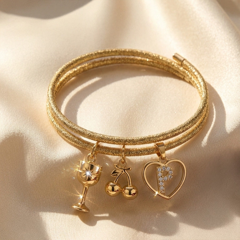 Gold-tone double-strand charm bracelet featuring a sparkling wine glass, cherry, and heart-shaped initial “P” charms, displayed on a soft satin background.