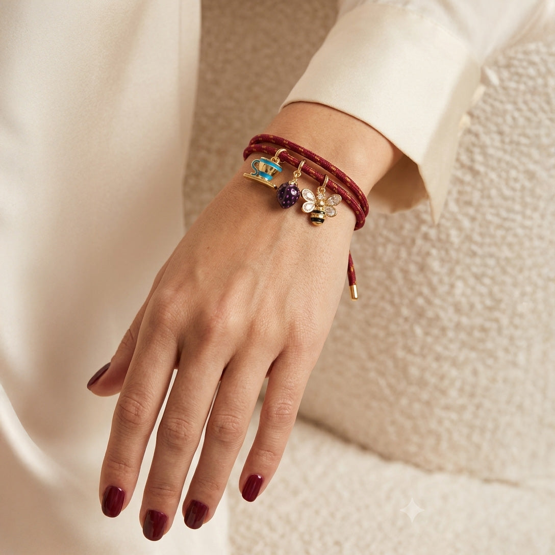 Gold-tone burgundy braided charm bracelet worn on the wrist, featuring a blue cup, purple grape, and sparkling bee charms, styled with a white outfit.