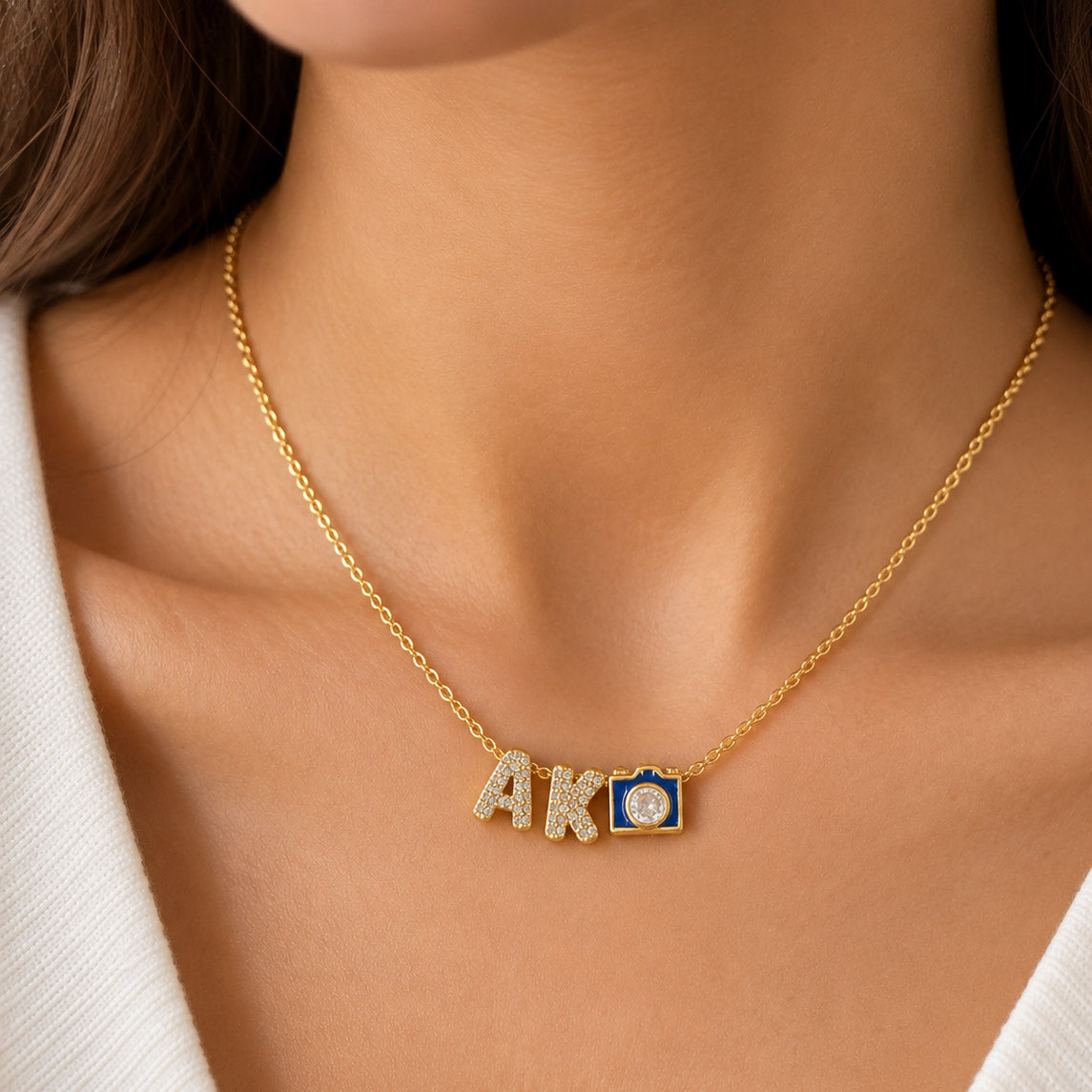 Close-up of a person's neck wearing a thin gold chain necklace with custom charms: two crystal-paved letters "A" and "K" followed by a small blue and gold camera pendant with a rhinestone lens. 
