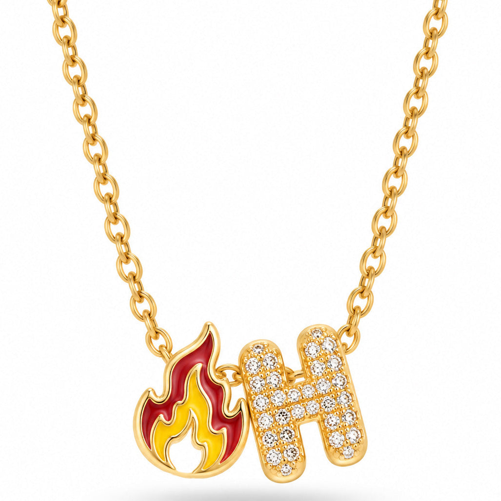 A studio shot of a delicate gold link necklace holding two charms: a colorful red and yellow flame pendant and a rhinestone-encrusted initial letter "H".