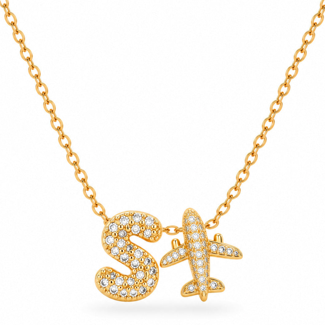 A studio shot of a thin gold link necklace holding two diamond-encrusted pendants: a puffy letter "S" and a small airplane.