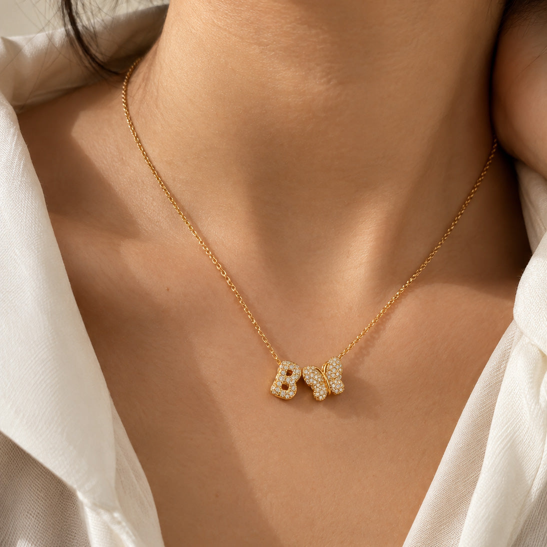 Close-up of a person's neck wearing a thin gold link necklace with custom sparkling charms: a rhinestone-encrusted letter "B" and a matching crystal butterfly pendant.