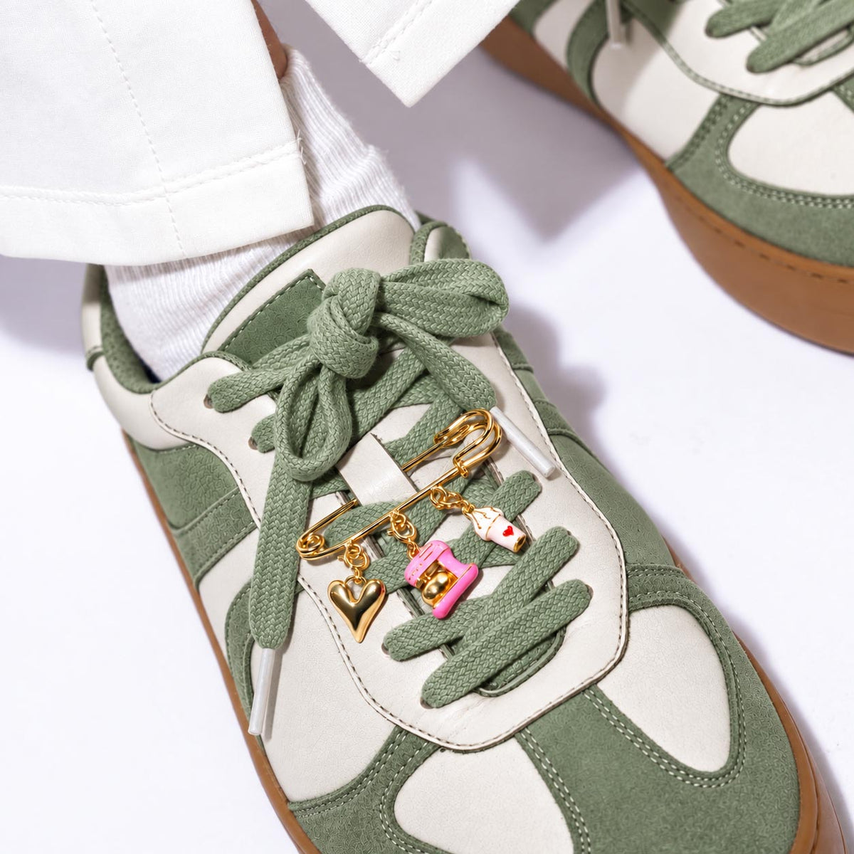 Gold-tone safety pin charm brooch attached to a green-and-white sneaker, featuring dangling charms including a heart, pink hand mixer, and small decorative charm.