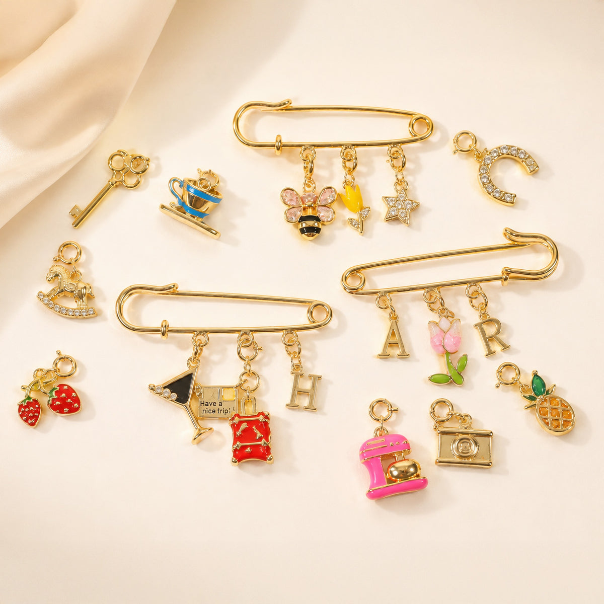 Gold-tone safety pin charm holder featuring interchangeable charms including initials, flowers, bee, star, cocktail glass, travel suitcase, camera, key, pineapple, strawberry and teacup, displayed on a soft beige background.