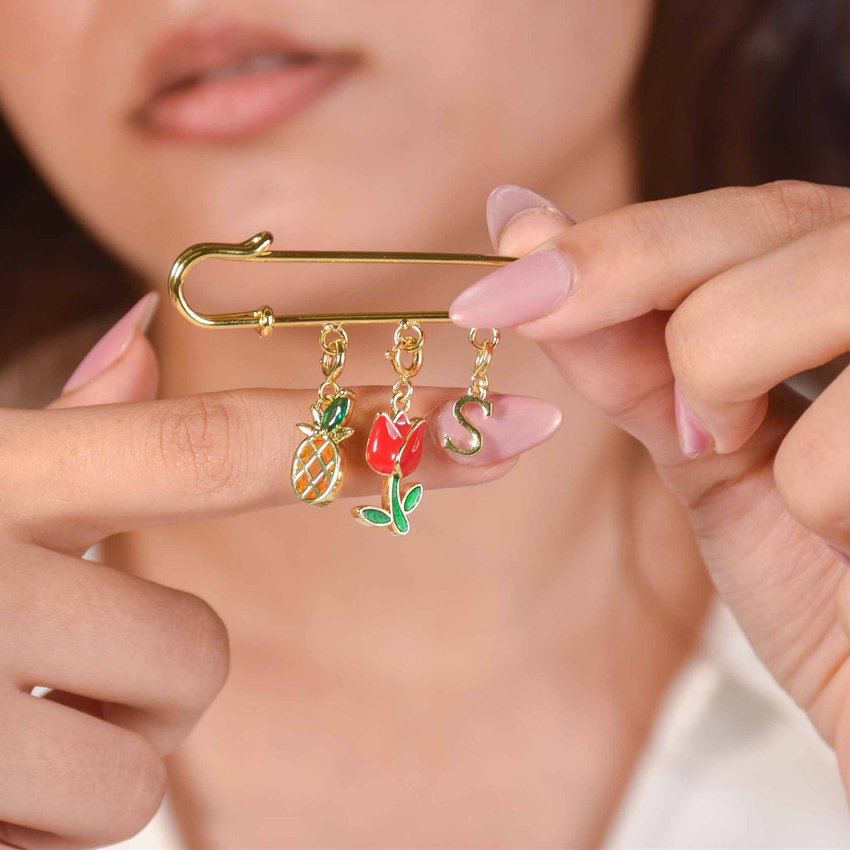 Gold-tone personalized safety pin charm holder with pineapple, red tulip, and initial “S” charms, held by hand against a white outfit.