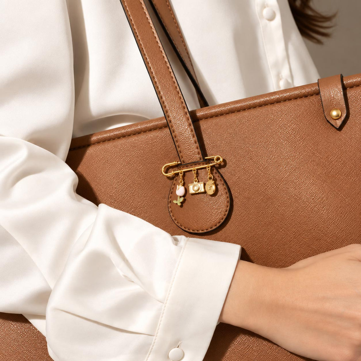 Gold-tone safety pin charm brooch attached to a brown handbag, featuring dangling decorative charms including a heart, camera, and other miniature charms.